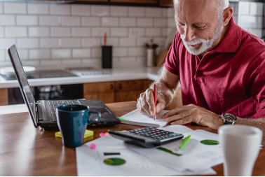 Older man at his kitchen table, writing notes on paper in front of a laptop.